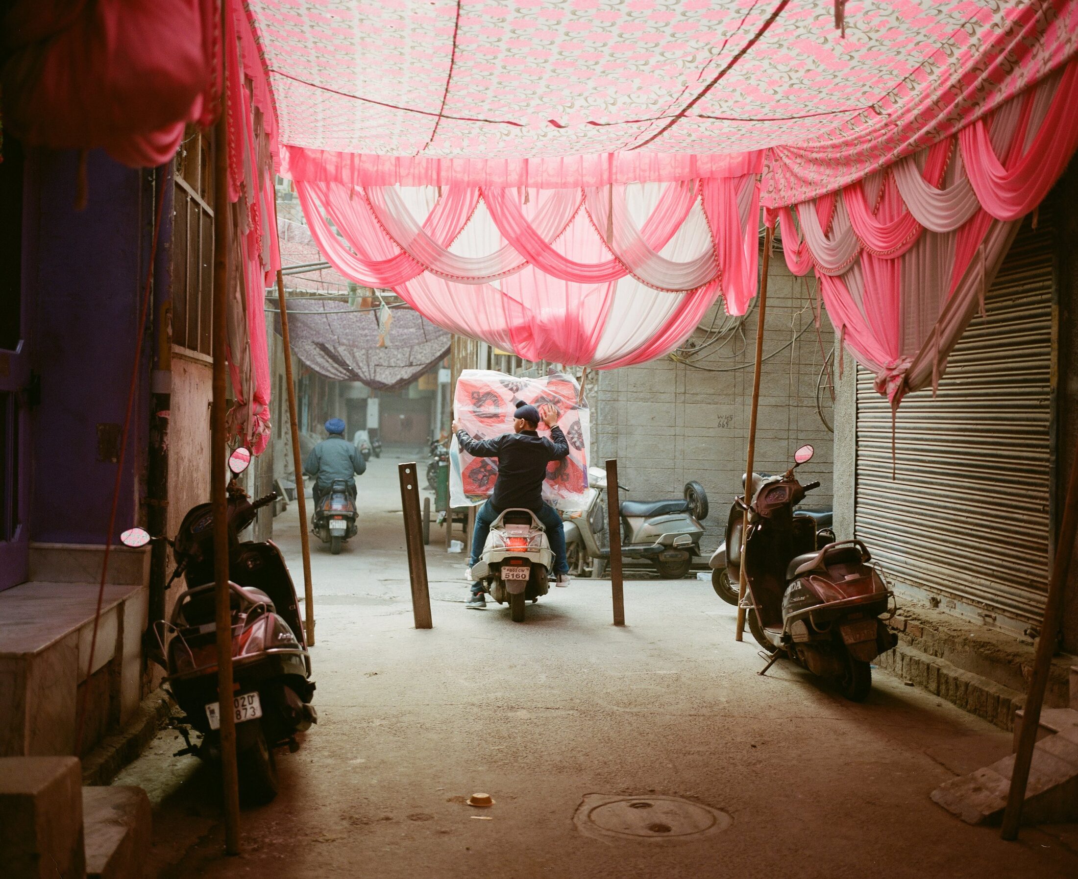 Pink wedding canopies strung above an alley of parked scooters, Punjab